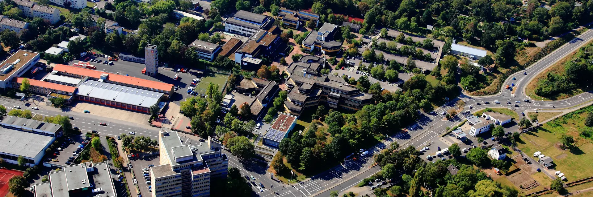 Luftaufnahme einer Stadt mit mehreren Gebäuden, Straßen und grünen Flächen. Im Hintergrund sind Sportanlagen und Wohngebäude sichtbar. Die Umgebung wirkt urban und gut bewaldet, mit lebhaftem Straßenverkehr und ausgeglichener Architektur.
