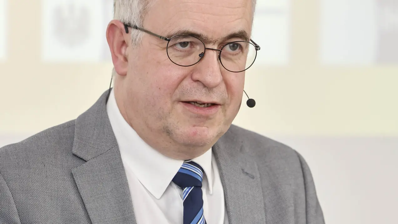 Ein Mann mit Brille und Anzug steht im Vordergrund, während er spricht. Er hat kurzes, graues Haar und trägt ein Hemd mit einer gestreiften Krawatte. Der Hintergrund ist unscharf und zeigt keine spezifischen Details.