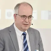 Ein Mann mit Brille und Anzug steht im Vordergrund, während er spricht. Er hat kurzes, graues Haar und trägt ein Hemd mit einer gestreiften Krawatte. Der Hintergrund ist unscharf und zeigt keine spezifischen Details.