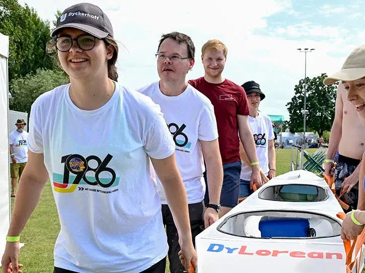 Eine Gruppe junger Menschen trägt ein Kajak über eine Wiese. Sie tragen T-Shirts mit dem Aufdruck "1986". Die Atmosphäre wirkt fröhlich und aktiv, während sie zusammenarbeiten, um das Kajak zu bewegen.