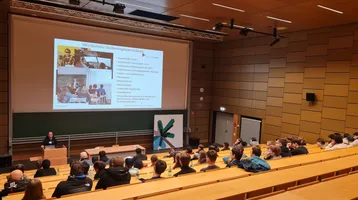 Ein Dozent steht an einem Podium in einem Hörsaal und präsentiert Informationen zu Studienmöglichkeiten. Im Hintergrund sind Folien mit Text und Bildern von Studierenden zu sehen. Das Publikum besteht aus zahlreichen Zuhörern, die aufmerksam dem Vortrag folgen.