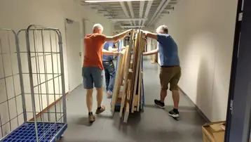 Drei Personen transportieren gemeinsam einen großen Stapel von Holzplatten durch einen langen Flur. Im Hintergrund sind Regale und Kisten sichtbar. Die Männer tragen kurze Hosen und arbeiten zusammen, um die Platten effektiv zu bewegen.