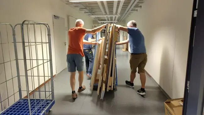 Drei Personen transportieren gemeinsam einen großen Stapel von Holzplatten durch einen langen Flur. Im Hintergrund sind Regale und Kisten sichtbar. Die Männer tragen kurze Hosen und arbeiten zusammen, um die Platten effektiv zu bewegen.