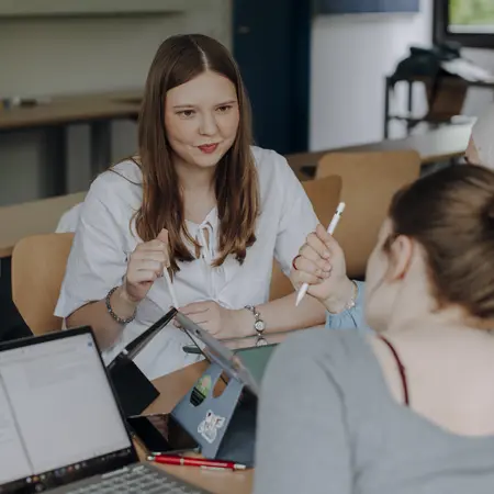 Man sieht drei Studierende im Vordergrund, die gemeinsam an einem Projekt arbeiten und lächeln.
