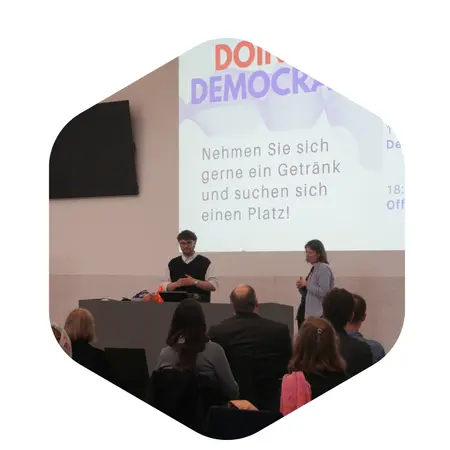 Ein Vortrag im Rahmen einer Veranstaltung zum Thema Demokratie. Eine Person steht am Rednerpult, während eine zweite Person auf der Bühne assistiert. Im Hintergrund sind Teilnehmende zu sehen, die sich in einem Raum umschauen. Ein Willkommenshinweis ist auf einer Leinwand zu sehen.