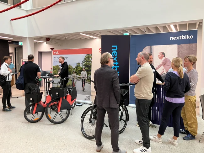 Eine Gruppe von Personen steht in einem modernen Innenraum, umgeben von Fahrrädern und Werbeflächen für den Mietrad-Service "nextbike". Die Leute scheinen sich zu unterhalten und Informationen auszutauschen. Im Hintergrund sind Plakate sichtbar, die den Service bewerben.