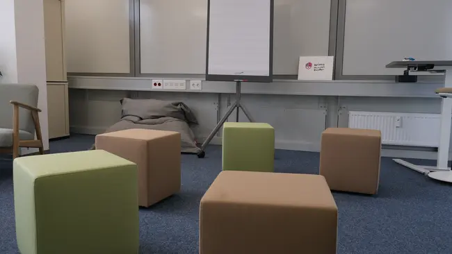 Der Raum ist mit mehreren Sitzwürfeln in verschiedenen Farben eingerichtet, darunter ein grüner und braune Exemplare. Ein Flipchart steht im Hintergrund, und eine beschriftete Tafel ist sichtbar. Der Boden ist mit Teppich ausgelegt.