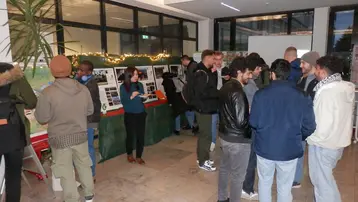 Eine Gruppe von Menschen steht in einem Raum, in dem verschiedene Informationsstände aufgestellt sind. Einige Personen unterhalten sich, während andere die ausgestellten Materialien ansehen. Der Raum hat große Fenster und eine freundliche Atmosphäre. Pflanzen sind ebenfalls im Hintergrund sichtbar.