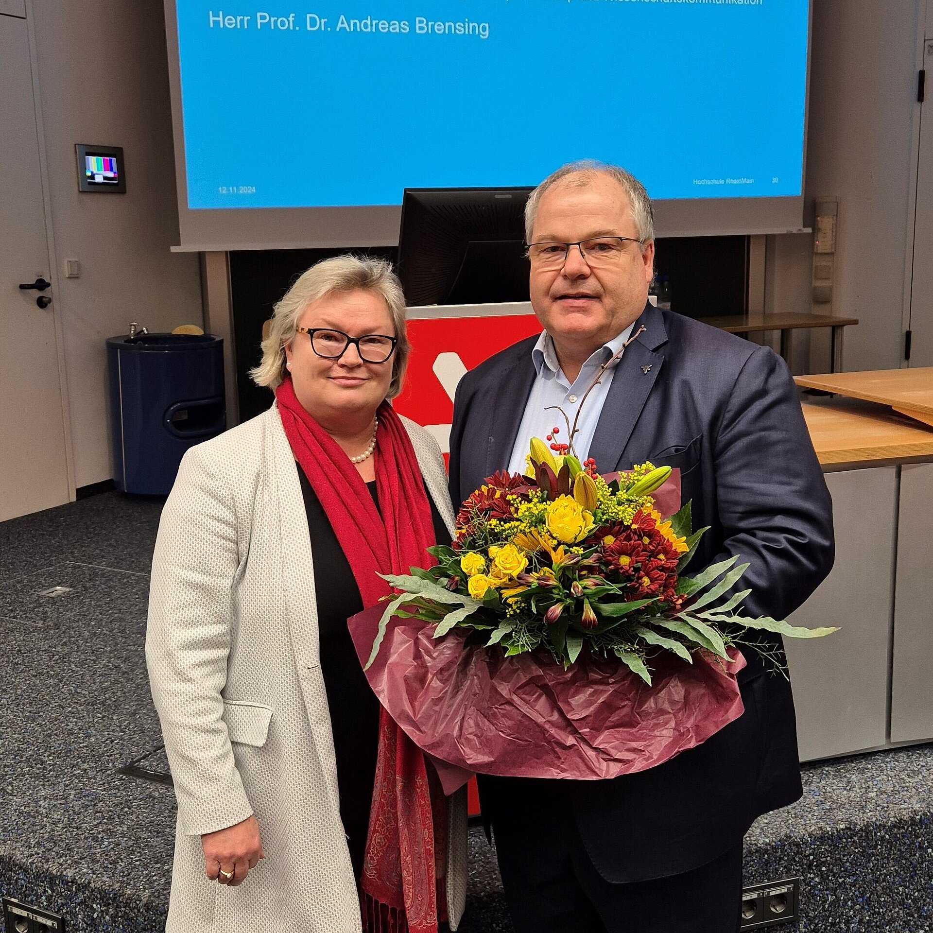 Prof. Dr. Eva Waller steht neben Prof. Dr. Andreas Brensing, der einen bunten Blumenstrauß in der Hand hält
