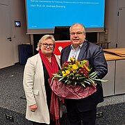 Prof. Dr. Eva Waller steht neben Prof. Dr. Andreas Brensing, der einen bunten Blumenstrauß in der Hand hält