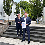 Prof. Dr. Eva Waller und Dr. Peter Adler stehen auf einer Treppe, er hält seine Ernennungsurkunde in der Hand