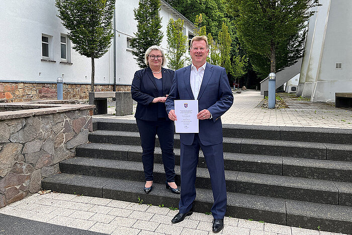 Prof. Dr. Eva Waller und Dr. Peter Adler stehen auf einer Treppe, er hält seine Ernennungsurkunde in der Hand