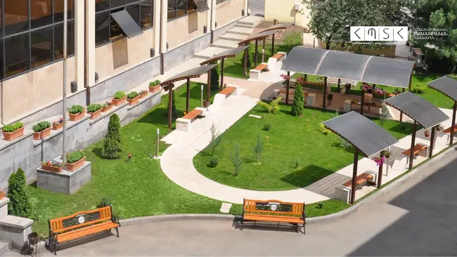 Eine grüne Gartenfläche mit Rasen und bepflanzten Beeten. In der Mitte führt ein geschwungener Weg zu einem überdachten Sitzbereich mit Holzbänken. Die Umgebung ist von einer modernen Architektur umgeben, die Schatten bietet und zum Verweilen einlädt.