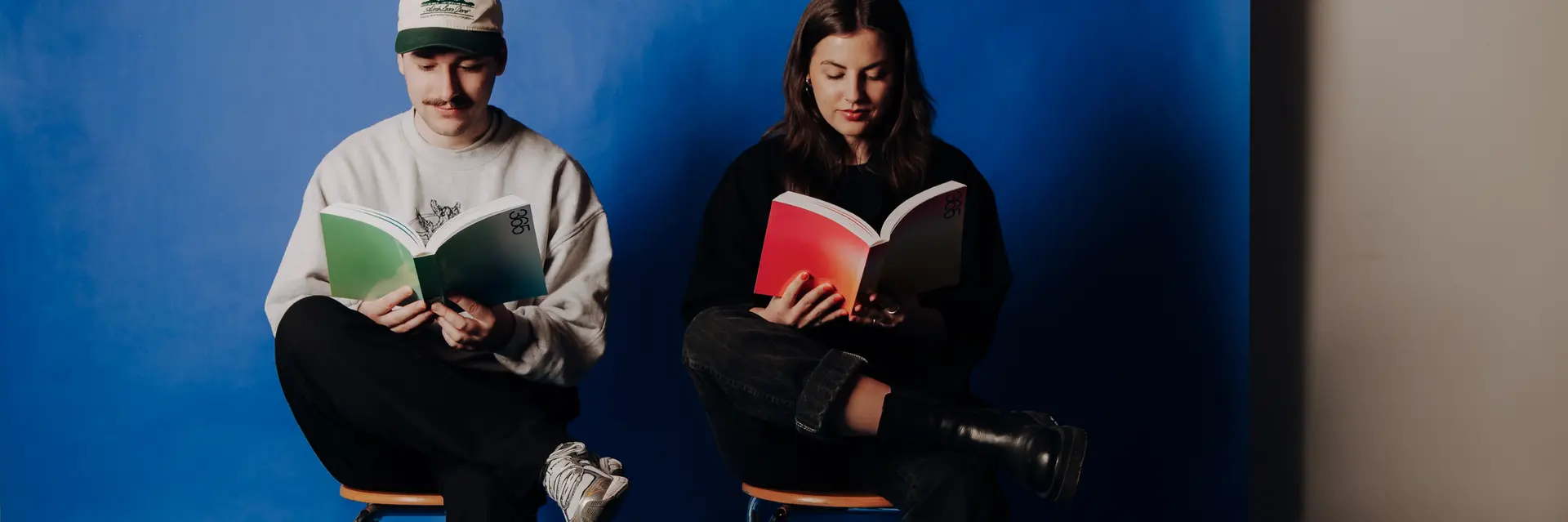 Zwei Personen sitzen auf Stühlen vor einem blauen Hintergrund. Die eine Person trägt eine Cap und hält ein grünes Buch, während die andere Person ein rotes Buch liest. Beide scheinen in ihre Lektüre vertieft zu sein.