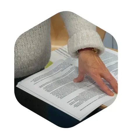 Eine Hand blättert durch ein offenes Buch mit bedruckten Seiten. Die Person trägt einen gestrickten Pullover und hat eine Armierung am Handgelenk.
