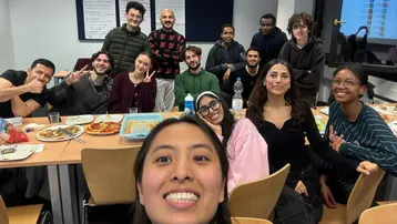 Eine Gruppe von 15 Personen sitzt an einem Tisch, umgeben von Tellern mit Essen und Getränken. Sie lächeln und posieren für ein Foto, während einige die Kamera direkt anschauen. Der Raum wirkt freundlich und gesellig. Die Stimmung ist entspannt und fröhlich.