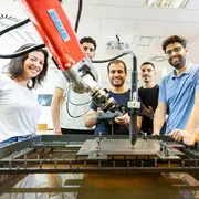 Junge Menschen stehen an einem roten Industrieroboter und schauen in die Kamera.