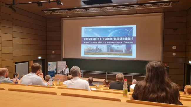 Das Bild zeigt eine Präsentation in einem Seminarraum. Auf der Leinwand steht der Titel „Wasserstoff als Zukunftstechnologie“ mit dem Untertitel „Internationale Märkte & innovative FTX-Lösungen“. Im Vordergrund sind Personen auf Sitzen zu sehen, die sich die Präsentation anschauen.