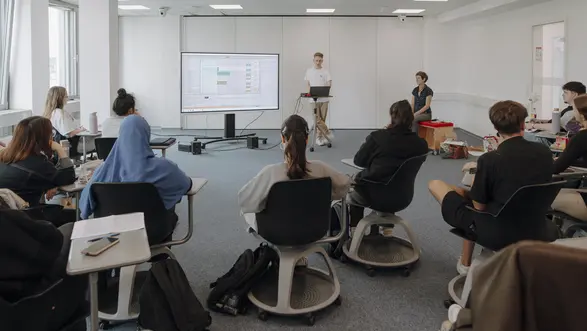 Man sieht viele Studierende, die alle in die gleiche Richtung schauen und einem Vortragenden zuhören. Er zeigt etwas auf einem großen Bildschirm mit dem er verbunden ist. Neben ihm sitzt eine Professorin und hört gespannt zu.