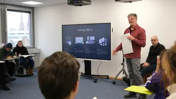 Ein Dozent steht vor einer Gruppe von Teilnehmern in einem Seminarraum und präsentiert Informationen auf einem Bildschirm. Auf dem Bildschirm ist ein Datum und weitere Inhalte sichtbar. Die Zuhörer sind aufmerksam und nehmen Notizen.