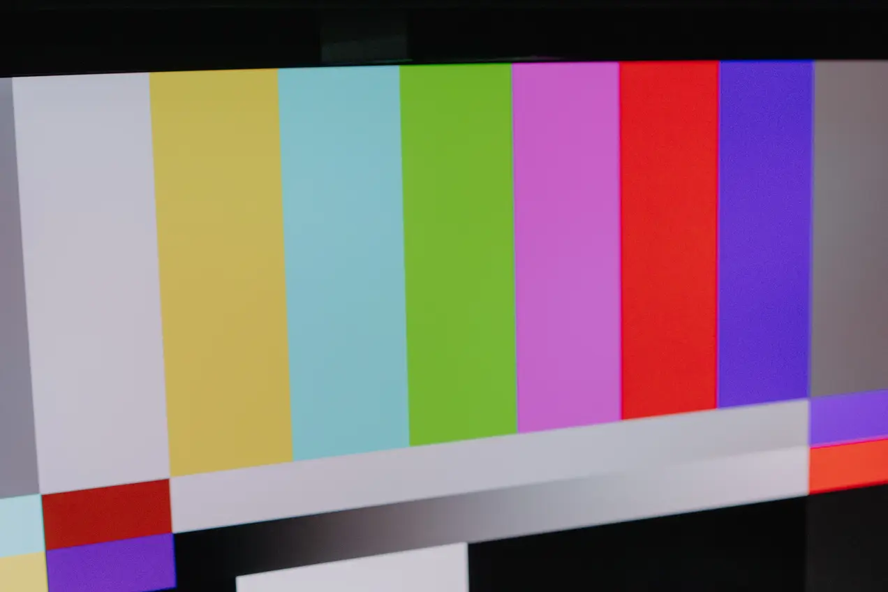 Ein Mischpult steht vor einem Monitor mit einem farbigen Testbild. Der Bildschirm zeigt vertikale Streifen in verschiedenen Farben. Das Mischpult hat mehrere Regler und Knöpfe, die zur Steuerung von Audio- oder Videosignalen verwendet werden können.