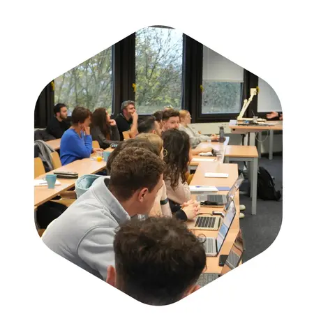 Eine Gruppe von Studierenden sitzt an Tischreihen in einem Klassenraum. Einige nutzen Laptops, während sie auf einen Vortragenden in der Nähe achten. Durch große Fenster ist Tageslicht zu sehen.