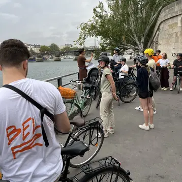 Eine Gruppe von Menschen steht an einem Flussufer, einige haben Fahrräder dabei. Eine Person spricht zu der Gruppe, während andere zuhören. Einige tragen Helme und wirken bereit für eine Radtour. Die Umgebung ist städtisch mit Bäumen im Hintergrund.