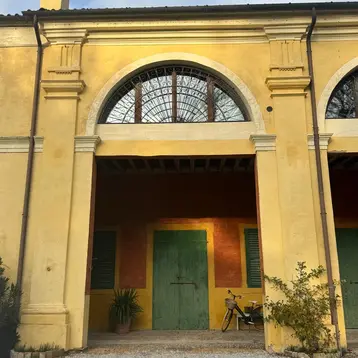 A historic building with a yellow facade and large archways. The doors are green, and there is a bicycle in the foreground. Plants can be seen surrounding the space. The sky is partly cloudy.