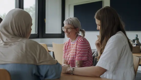 Man sieht drei Studierende im Vordergrund, die gemeinsam an einem Projekt arbeiten und lächeln.