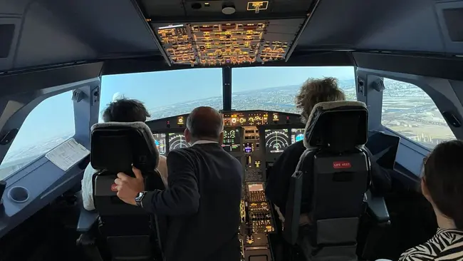 Im Flugsimulator eines A320 sitzen drei Personen, die auf das Cockpit-Display blicken. Die linke Person steuert das Flugzeug, während die anderen Personen ihm zuschauen. 