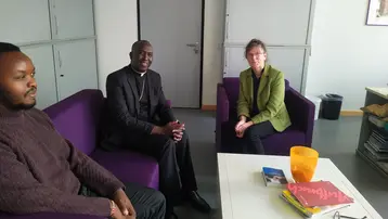 Drei Personen sitzen in einem modernen Raum auf lila Sofas. Ein Mann in einem schwarzen Anzug und zwei weitere Personen in lässiger Kleidung unterhalten sich. Auf dem Tisch neben ihnen liegen Bücher und eine orangefarbene Schüssel. Die Atmosphäre wirkt freundlich und entspannt.