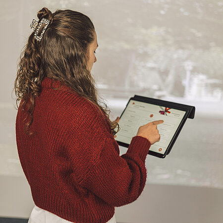 Frau von hinten mit Tablet Man sieht eine Frau, die den Rücken zu uns gewandt hat und verschiedene Buttons auf dem Tablet drückt. Der Hintergrund ist ein Spielplatz auf der Leinwand.