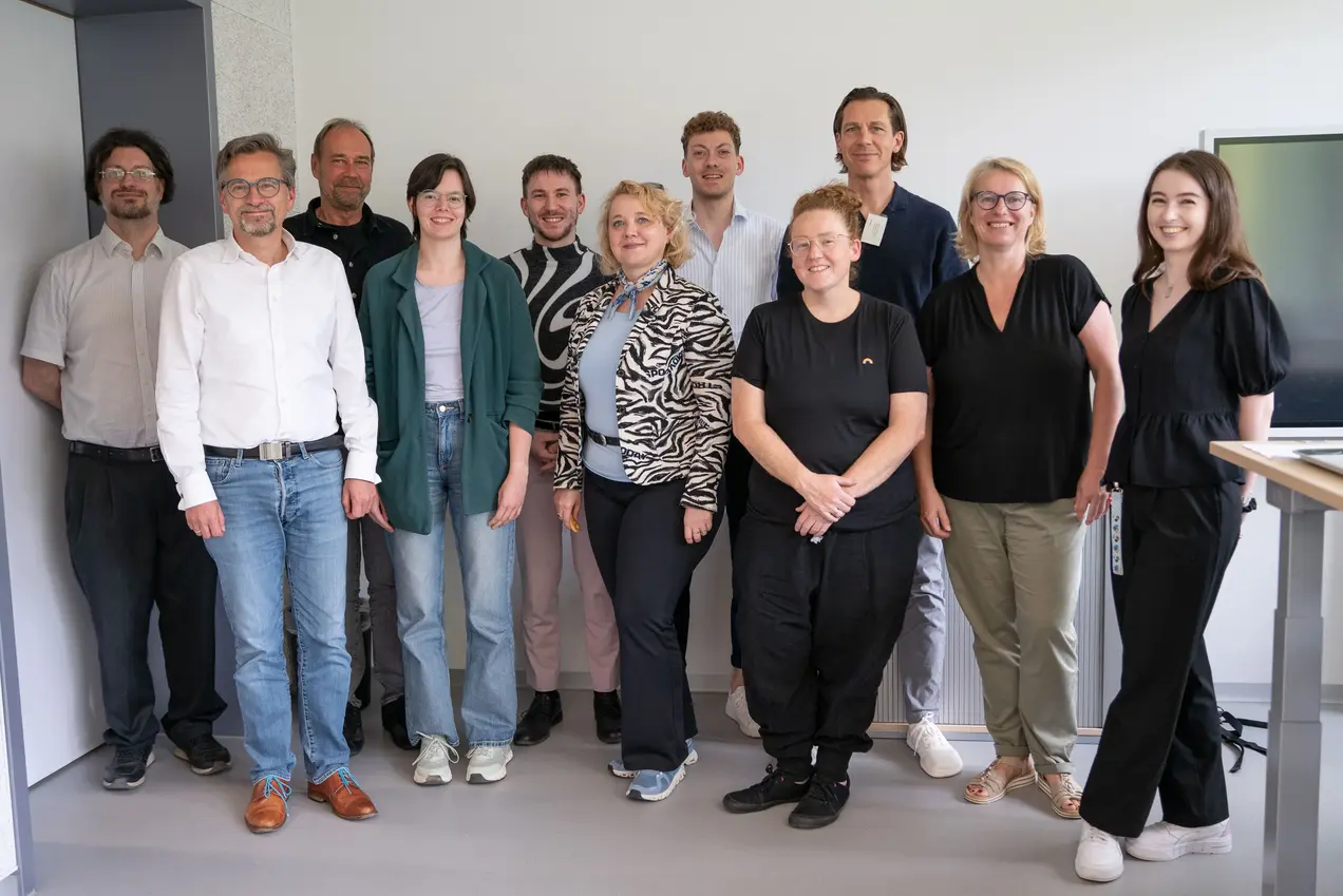 Eine Gruppe von zwölf Personen steht in einem modernen Raum. Sie sind vielfältig gekleidet und lächeln in die Kamera. Im Hintergrund sind Wände und ein Bildschirm zu sehen, die den Raum gestalten. Die Atmosphäre wirkt freundlich und einladend.