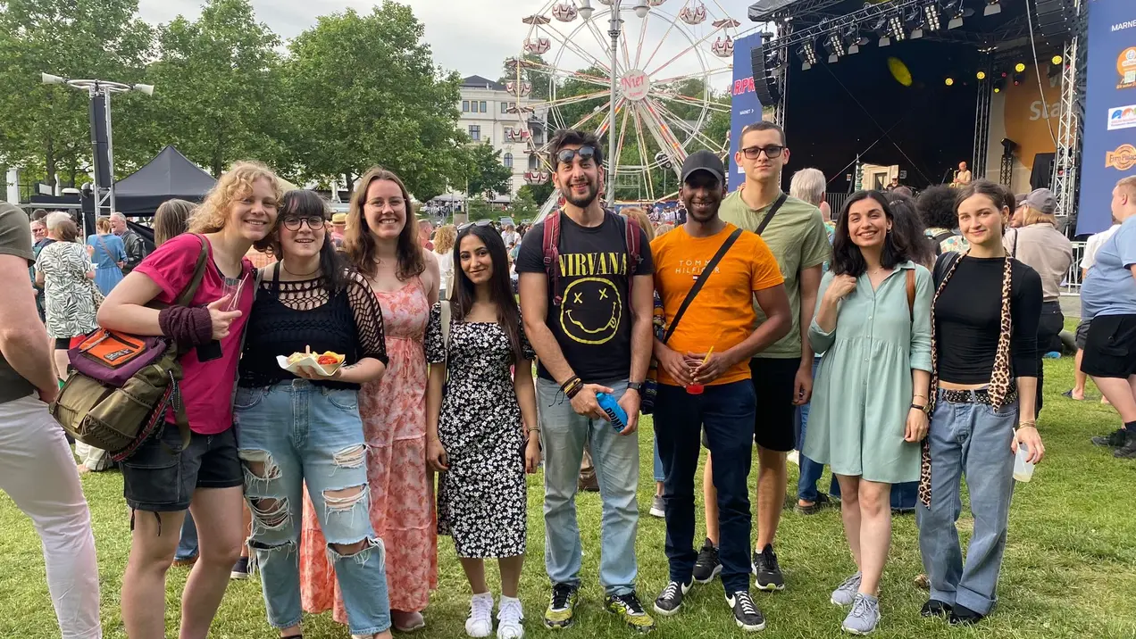 Gruppendfoto von acht Personen, die lächeln und zusammenstehen. Sie tragen verschiedene Sommerkleidungsstile und befinden sich auf einem Festivalgelände mit Bühnen im Hintergrund. Eine riesige Radattraktion ist ebenfalls sichtbar, umgeben von grünen Bäumen und weiteren Festivalbesuchern.