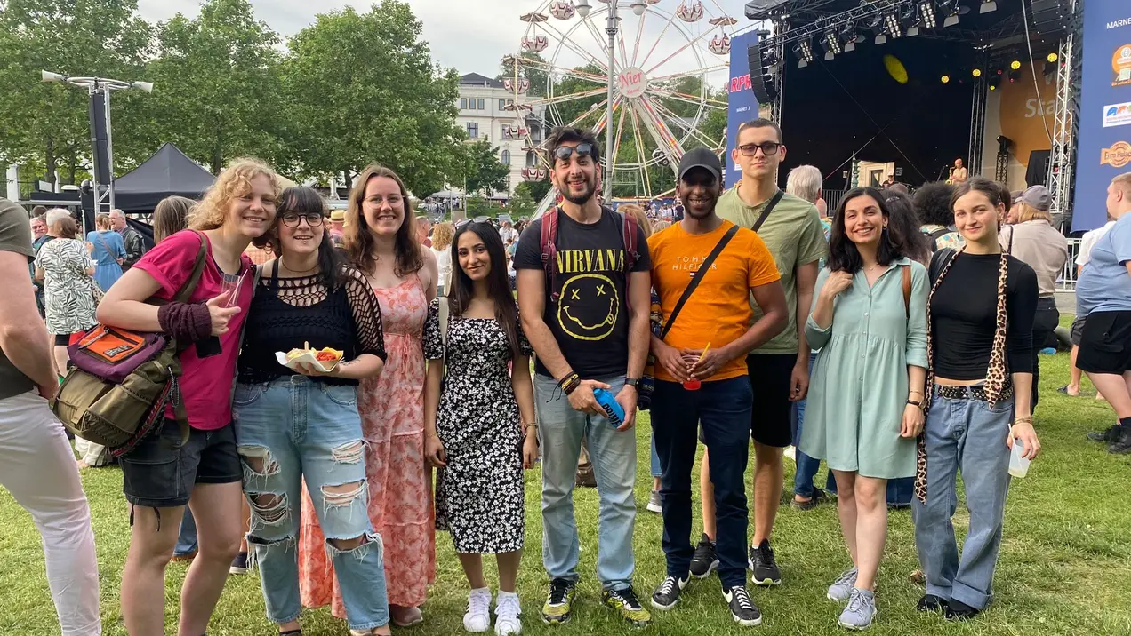 A group of nine friends stands together at an outdoor festival, smiling and posing for the photo. They are dressed in casual summer outfits, surrounded by a lively crowd and a large ferris wheel in the background. The atmosphere appears joyful and festive.
