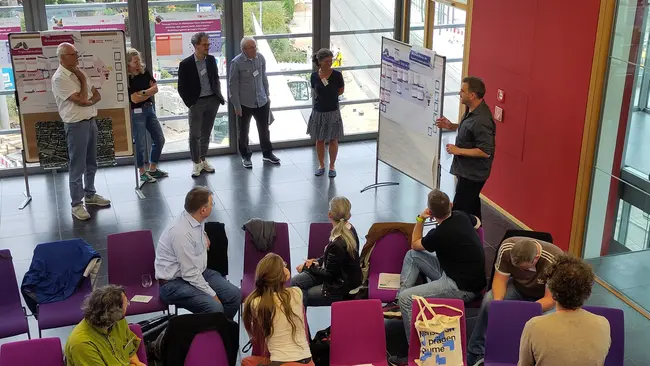 Eine Gruppe von Personen nimmt an einer Präsentation oder Besprechung teil. Ein Mann steht vor einem Flipchart und erklärt etwas, während andere Teilnehmer aufmerksam zuhören. Die Szene spielt sich in einem modernen Raum mit großen Fenstern ab, wodurch Licht und Ausblick sichtbar sind.