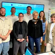 Eine Gruppe von neun Personen steht vor einem Bildschirm, auf dem „Herzlich Willkommen zur Jugendsitzung“ steht. Die Teilnehmer wirken freundlich und engagiert, einige halten Stifte oder Notizen in der Hand. Der Raum ist hell und kreativ gestaltet.
