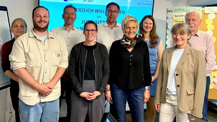 Eine Gruppe von neun Personen steht vor einem Bildschirm, auf dem „Herzlich Willkommen zur Jugendsitzung“ steht. Die Teilnehmer wirken freundlich und engagiert, einige halten Stifte oder Notizen in der Hand. Der Raum ist hell und kreativ gestaltet.