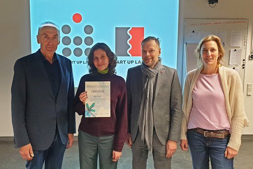 Prof. Rüdiger Pichler steht mit der Gewinnerin Lara Čadež sowie Dr. Klaus Bernsau und Dr. Sandra Steinbrink vom Projekt RheinMain StartUpLabs vor dem Siegerlogo