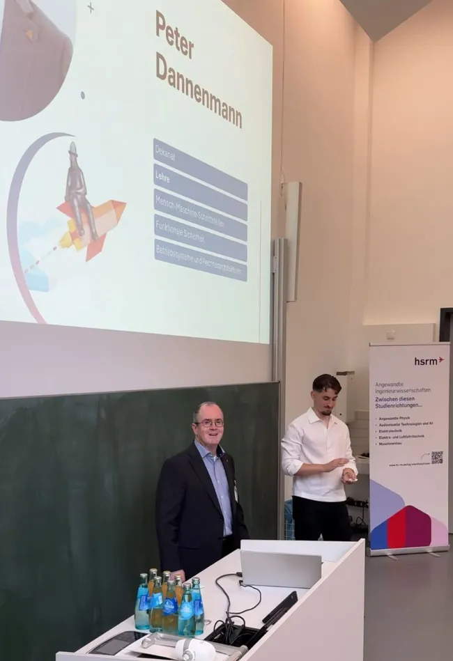 Eine Präsentation findet in einem Seminarraum statt. Zwei Männer stehen vor einer Leinwand mit dem Namen "Peter Dannenmann" und Informationen über ihn. Auf einem Tisch sind Wasserflaschen platziert. Im Hintergrund steht ein Banner der Hochschule.