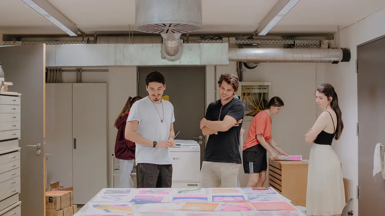 Design-Studierende betrachten sich Entwürfe auf einem Tisch am Campus unter den Eichen