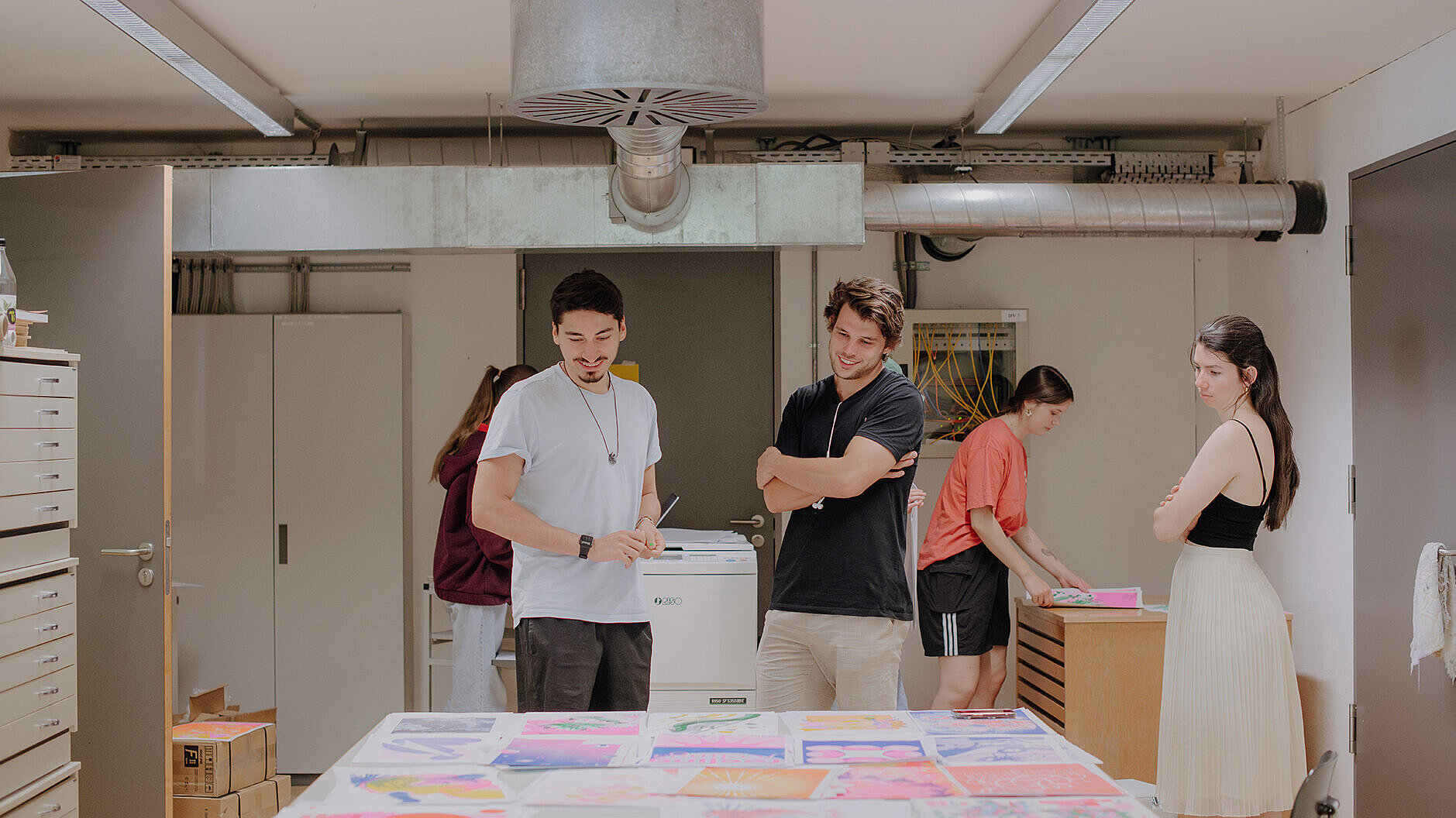 Design-Studierende betrachten sich Entwürfe auf einem Tisch am Campus unter den Eichen