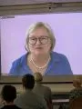 Eine Frau mit Brille und blauer Bluse spricht auf einer Videoleinwand. Im Vordergrund sind unscharf einige Zuhörer zu sehen, die der Präsentation aufmerksam folgen. Der Hintergrund ist einfarbig und schlicht.