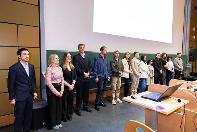 Eine Gruppe von mehreren Personen steht in einem Seminarraum aufgereiht. Einige tragen formelle Kleidung, andere legere Outfits. Im Vordergrund ist ein Tisch mit einem Laptop und technischem Equipment zu sehen. Im Hintergrund ist eine Leinwand, die leer ist.