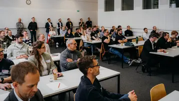 Eine Gruppe von etwa fünfzig Menschen sitzt an Tischen in einem Seminarraum. Die Teilnehmer hören aufmerksam zu, einige machen Notizen. Auf der Wand ist eine Uhr zu sehen, die die Zeit anzeigt. Das Ambiente ist konzentriert und professionell.