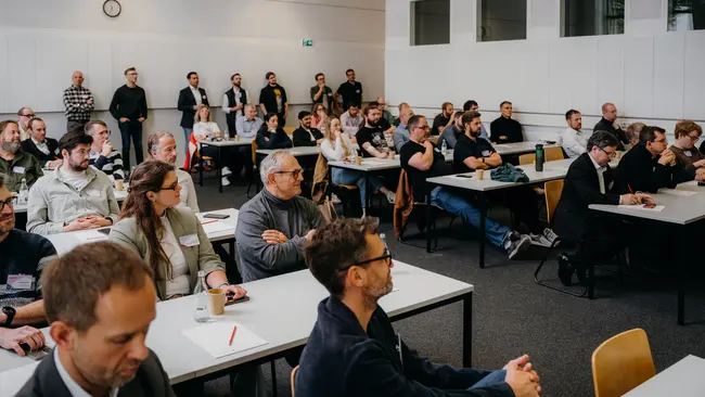 Eine Gruppe von etwa fünfzig Menschen sitzt an Tischen in einem Seminarraum. Die Teilnehmer hören aufmerksam zu, einige machen Notizen. Auf der Wand ist eine Uhr zu sehen, die die Zeit anzeigt. Das Ambiente ist konzentriert und professionell.