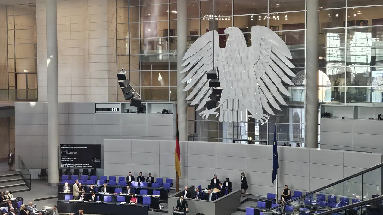 Im Bild ist der Bundestag mit dem Reichstagsgebäude zu sehen. Im Vordergrund befinden sich Abgeordnete in einem Sitzungssaal, während im Hintergrund der große deutsche Bundesadler an der Wand prangt. Die Atmosphäre wirkt formal und konzentriert.