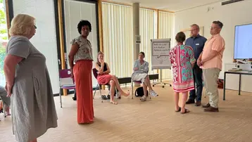 Eine Gruppe von sieben Personen steht in einem hellen Raum, viele schauten sich an und diskutieren. Einige sind in einem informellen Setting mit Stühlen und einem Flipchart. Die Atmosphäre wirkt interaktiv und gemeinschaftlich, wahrscheinlich handelt es sich um ein Meeting oder einen Workshop.