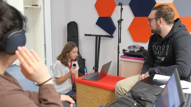 Eine junge Frau sitzt auf einem Hocker und spricht in ein Mikrofon, während sie auf einen Laptop schaut. Ein Mann neben ihr hört aufmerksam zu. Eine weitere Person, die Kopfhörer trägt, ist im Hintergrund zu sehen. Die Umgebung ist modern und freundlich gestaltet.