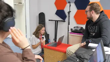 Eine junge Frau sitzt auf einem Hocker und spricht in ein Mikrofon, während sie auf einen Laptop schaut. Ein Mann neben ihr hört aufmerksam zu. Eine weitere Person, die Kopfhörer trägt, ist im Hintergrund zu sehen. Die Umgebung ist modern und freundlich gestaltet.